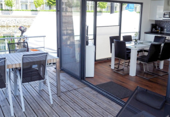 Zicht vanaf een woonboot met houten terras, buitentafel en moderne eethoek met keuken binnenin.