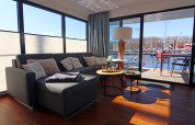 Salón moderno en casa flotante con sofá esquinal, mesas bajas, vistas al agua y terraza al fondo.