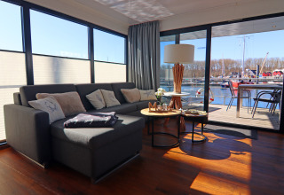Moderne husbåd-stue med stor sofa, sofaborde, udsigt til terrasse og havn gennem store glasdøre.