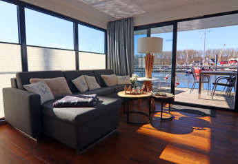 Salón moderno en casa flotante con sofá esquinal, mesas bajas, vistas al agua y terraza al fondo.