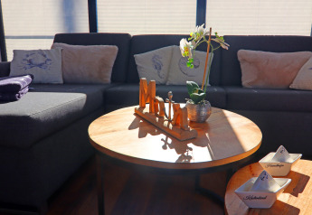 Sala de estar en casa flotante con sofá gris, cojines, flor y luz solar sobre una mesa de madera.