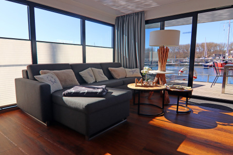 Bright living room on a houseboat with large sofa, coffee tables, and marina views through large windows in Germany.