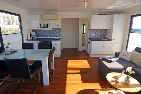 Bright, modern houseboat interior with dining table, sofa, open kitchen, and large windows with sunlight.