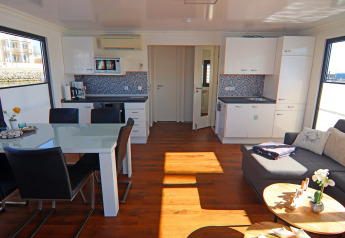 Interior luminoso y moderno de casa flotante con mesa de comedor, cocina abierta, sofá y grandes ventanas.