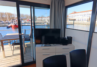 Living area of a houseboat with a TV, dining table, and marina view through large windows and deck.