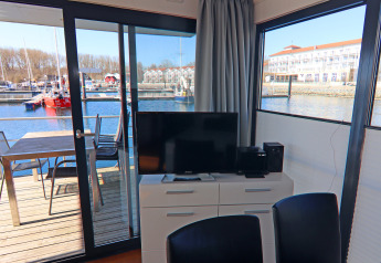 Soggiorno su una houseboat con TV, tavolo da pranzo e vista sul porto attraverso ampie finestre.