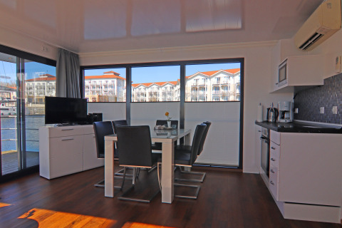 Modern houseboat interior featuring kitchen, dining area, and large windows overlooking a scenic marina.
