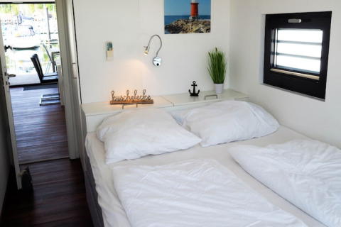 Cozy houseboat bedroom with white bedding, plant decoration, anchor, and a view to the dock outside.