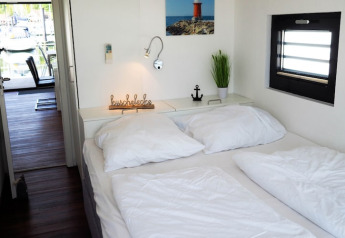 Dormitorio de casa flotante con ropa de cama blanca, decoración de planta y ventana con vista al muelle.