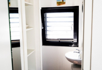 Baño luminoso en una casa flotante con ventana enmarcada en negro, estantes blancos y pequeño lavabo.