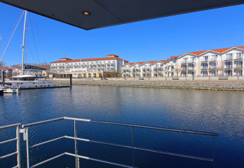 Vista desde una casa flotante hacia un puerto deportivo con veleros y apartamentos modernos al fondo.
