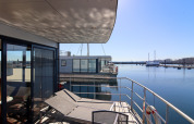 Casa flotante con terraza y tumbonas junto a un puerto tranquilo bajo un cielo despejado y soleado.