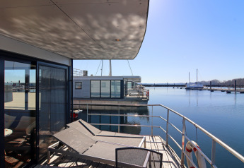Casa flotante con terraza y tumbonas junto a un puerto tranquilo bajo un cielo despejado y soleado.