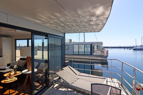 Modern houseboat with terrace and sun loungers on calm water, stylish interiors, and large sliding glass doors.