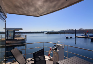 View from the houseboat terrace overlooking a calm harbor with chairs, lifebuoy, and steering post.