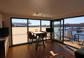 Sala de estar en el Houseboat Family & Friends con terraza y vistas al agua en Alemania, día soleado.