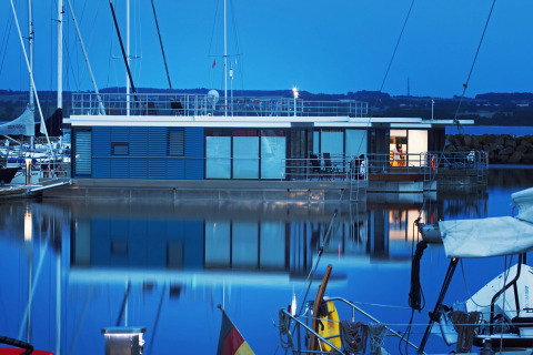 Modern houseboat 'Family & Friends mit Hund LP 10' docked in a German marina at dusk, softly illuminated.