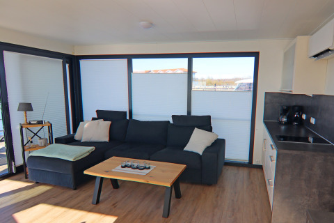Moderne stue på en husbåd i Tyskland med mørk sofa, sofabord, tekøkken og store vinduer.