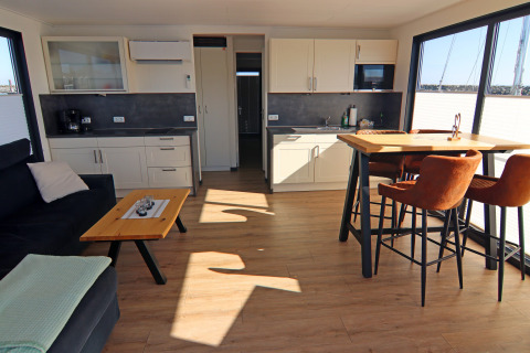 Modern houseboat interior with open kitchen, dining table, sofa and large windows overlooking the water.