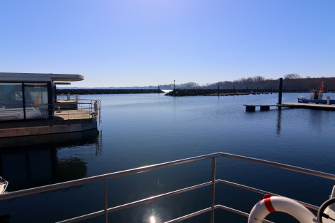 View from a houseboat over calm water, railings in foreground, pier and boats in distance, clear sky.