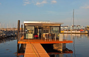 Een modern woonboot met glazen wanden en houten terras ligt aangemeerd in een zonnige jachthaven.