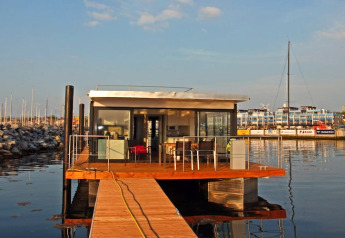 A modern houseboat with glass walls and a wooden deck is docked at a marina on a sunny day.