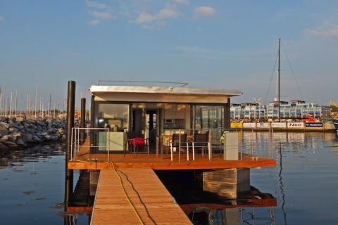 Une maison flottante moderne avec murs en verre et terrasse en bois amarrée dans un port au soleil.