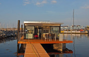 A modern houseboat with glass walls and a wooden deck is docked at a marina on a sunny day.