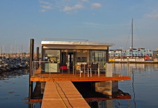 Une maison flottante moderne avec murs en verre et terrasse en bois amarrée dans un port au soleil.
