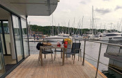 Terras van woonboot met tafel en stoelen, uitzicht op een jachthaven met zeilboten en groene heuvels.
