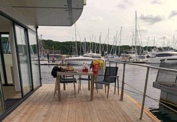 Terras van woonboot met tafel en stoelen, uitzicht op een jachthaven met zeilboten en groene heuvels.