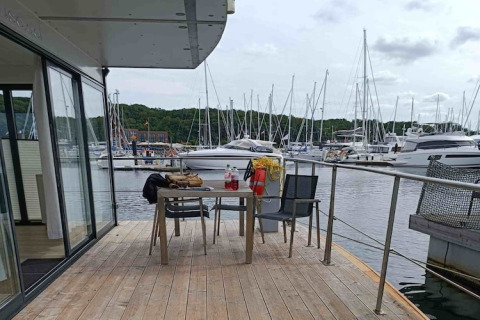 Terrasse de péniche avec table et chaises, vue sur un port de plaisance rempli de yachts et collines vertes.