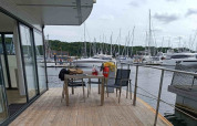 Houseboat deck with table and chairs, overlooking a marina filled with yachts and green forested hills.