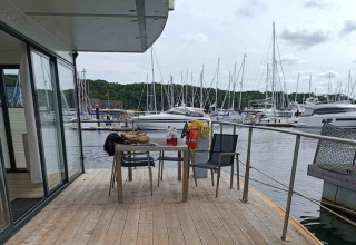 Terrasse de péniche avec table et chaises, vue sur un port de plaisance rempli de yachts et collines vertes.