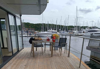 Terras van woonboot met tafel en stoelen, uitzicht op een jachthaven met zeilboten en groene heuvels.