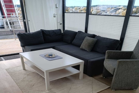Salon moderne sur une péniche avec canapé d’angle gris foncé, fauteuil et table basse blanche près des fenêtres.