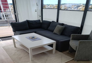 Salon moderne sur une péniche avec canapé d’angle gris foncé, fauteuil et table basse blanche près des fenêtres.