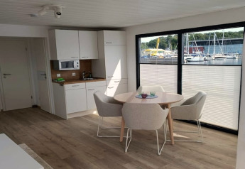 Modern houseboat interior with kitchenette and dining area, large windows offering marina views outside.