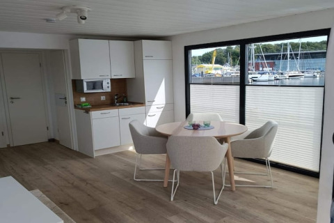 Intérieur de péniche moderne avec coin cuisine, salle à manger et grandes fenêtres donnant sur le port.