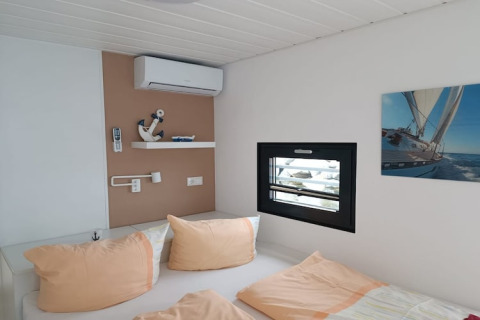 Chambre moderne sur une péniche avec fenêtre, tableau de voilier et oreillers pastel clairs.