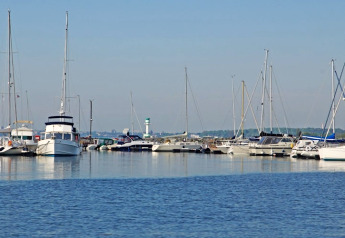 View of a marina with houseboats and sailboats docked, calm water and clear blue sky in the background.