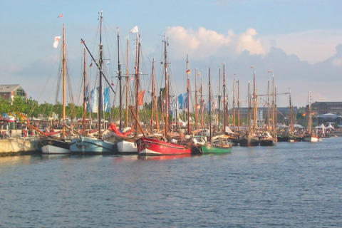Photo de voiliers colorés amarrés au port lors d’un festival animé, foule et drapeaux en arrière-plan.