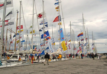 Bootshow met vele zeilboten en kleurrijke vlaggen in de haven, mensen wandelen over de kade onder wolken.