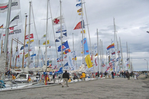 Salon nautique avec de nombreux voiliers et drapeaux colorés au port, des gens marchent sur le quai.