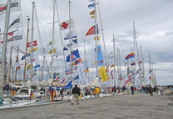 Boat show with numerous sailboats and colorful flags in harbor, people walking along the pier under clouds.