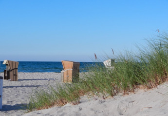 Strandscène met duingras en strandstoelen aan het zand, uitkijkend op de rustige blauwe zee en lucht.