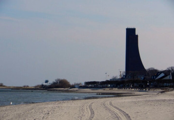 Zandstrand met een aparte hoge toren, rustig water en gebouwen bij een locatie voor woonboten.