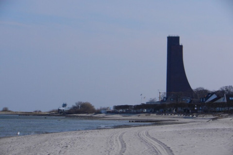 Plage de sable avec une tour haute originale, eau calme et maisons proches d’un site de péniche.