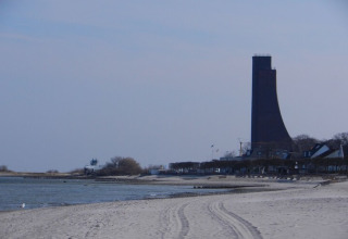 Plage de sable avec une tour haute originale, eau calme et maisons proches d’un site de péniche.