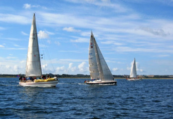 Meerdere zeilboten met bemanning racen over open water onder een deels bewolkte blauwe lucht nabij de kust.